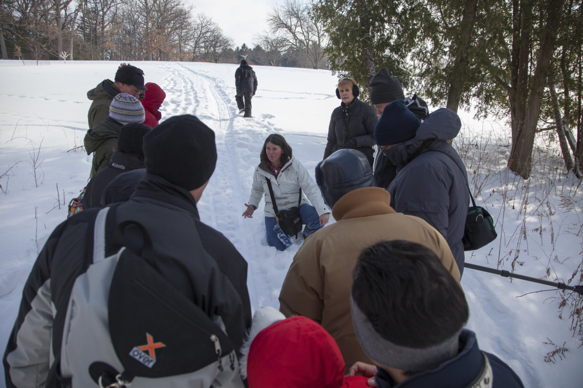 A group gathers around a teacher pointing out animal tracks in the snow.