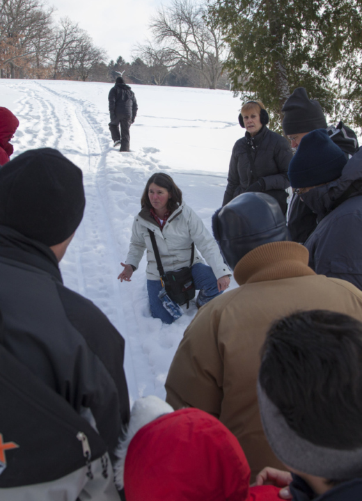 A group gathers around a teacher pointing out animal tracks in the snow.