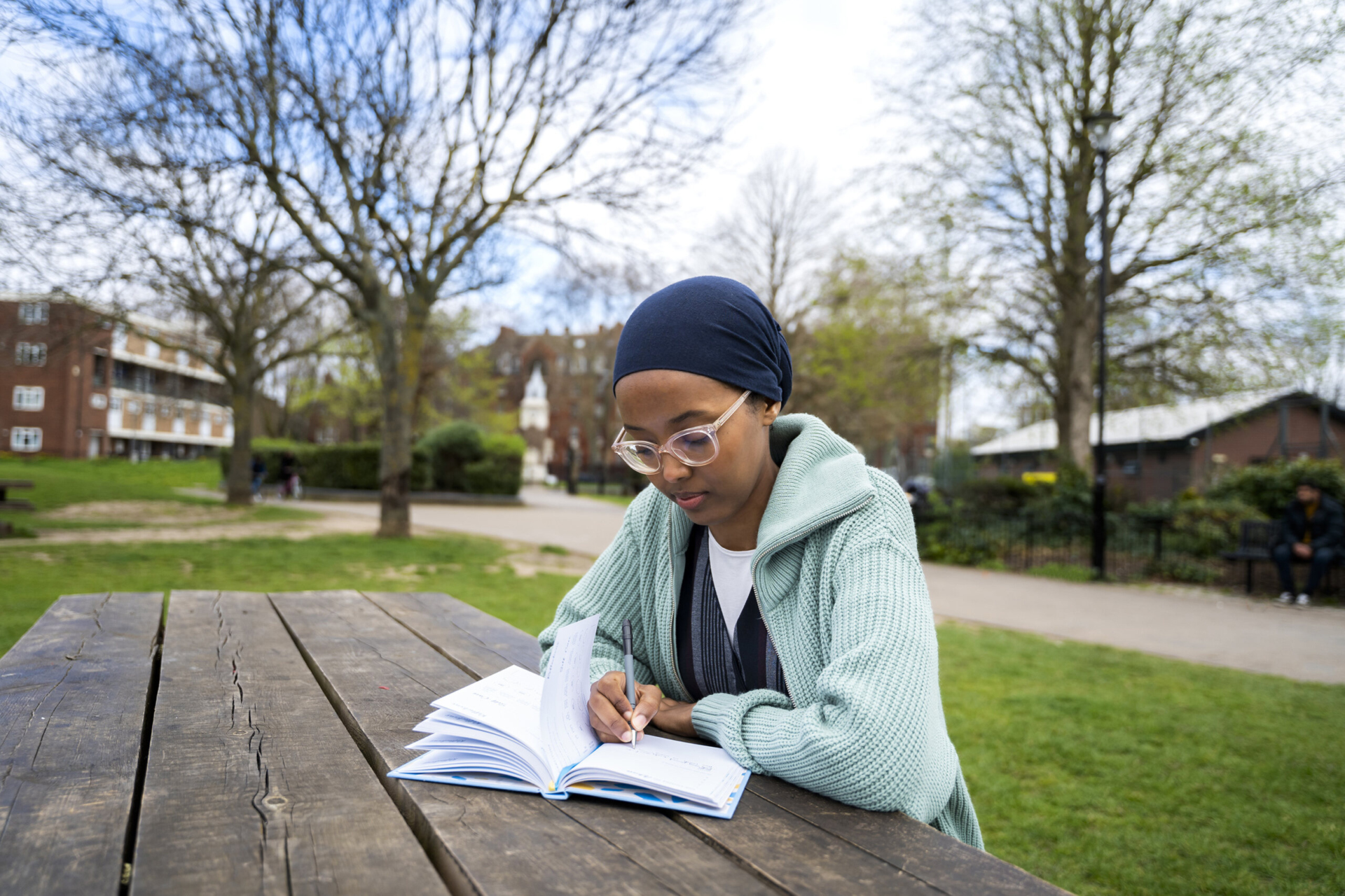 A young woman wearing a head scarf journaling while sitting at an outdoor picnic table.