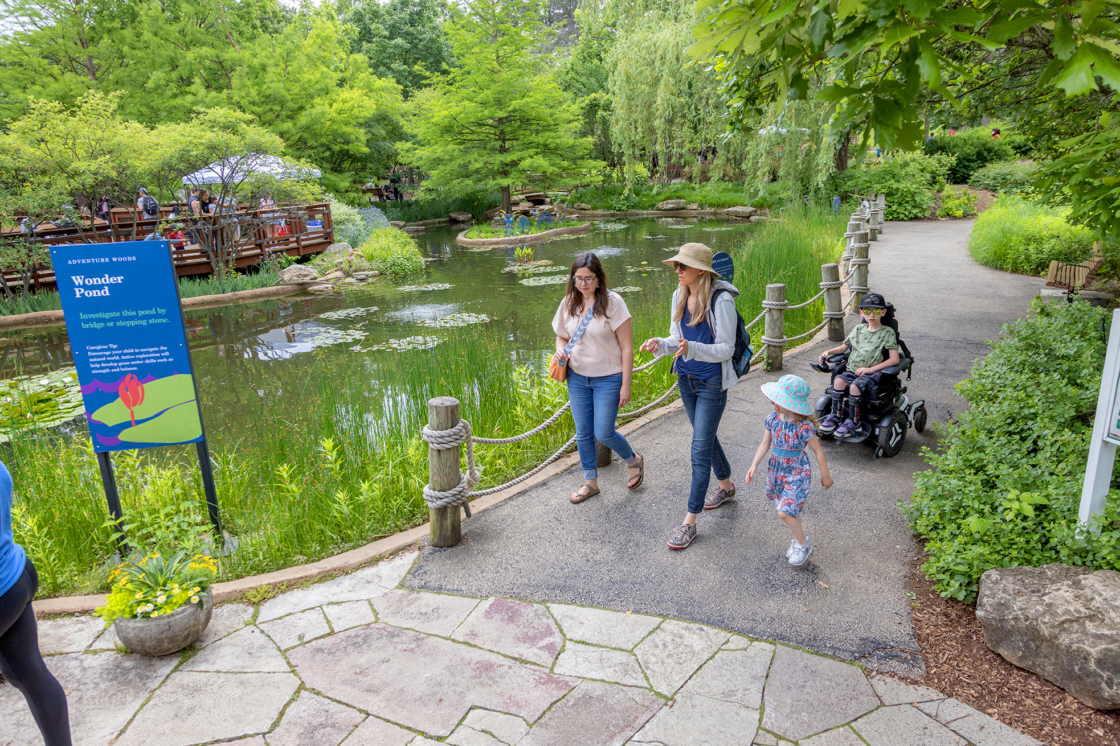 A family with a child in a motorized wheelchair stroll through the accessible Children's Garden near Wonder Pond.