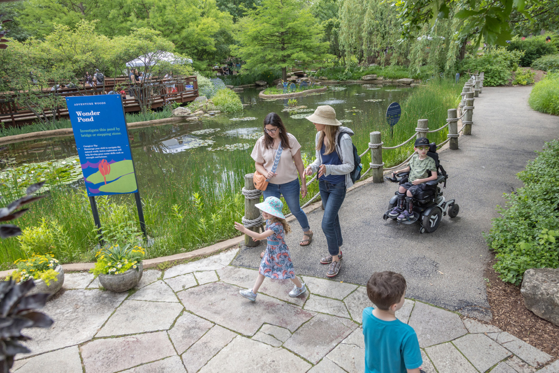 A family and a child in a motorized wheel chair make their way through the Children's Garden near Wonder Pond