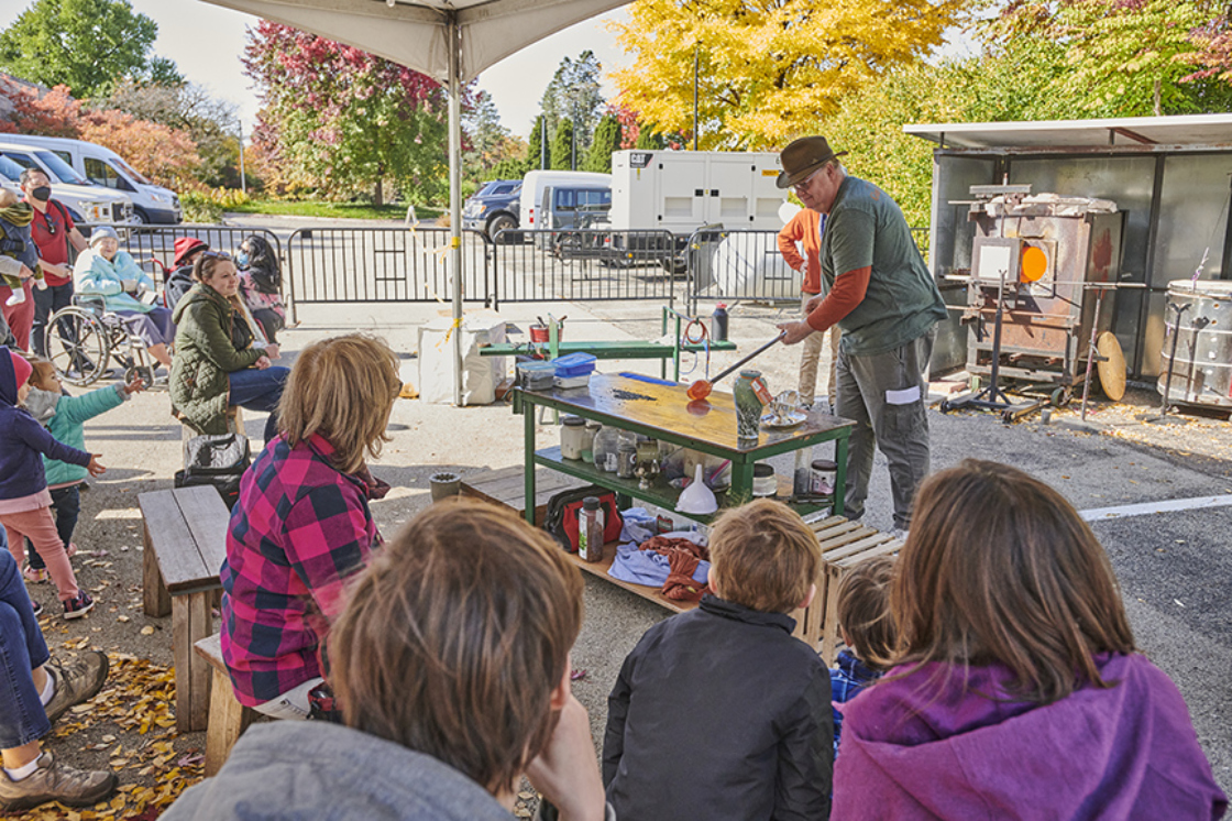 A glass artist giving a demonstration to a small crowd under a tent.
