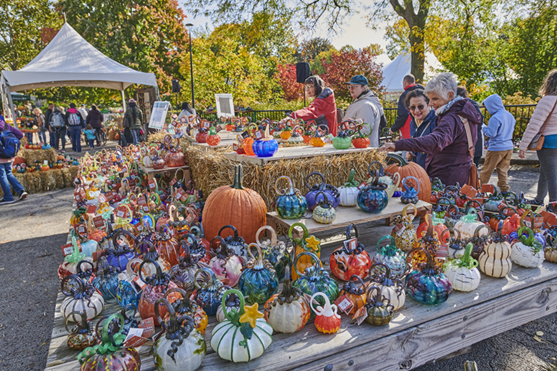 A group of guests shop at a large table full of glass pumpkins on a fall day.