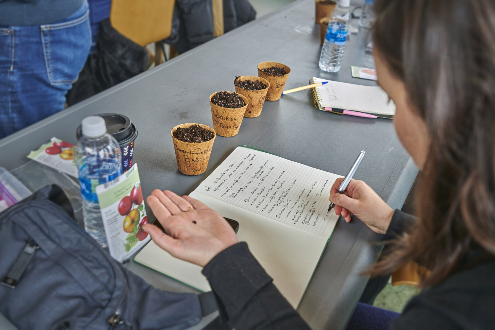 A person holding a seed and recording information in a notebook, four small seed starter pots on the table.