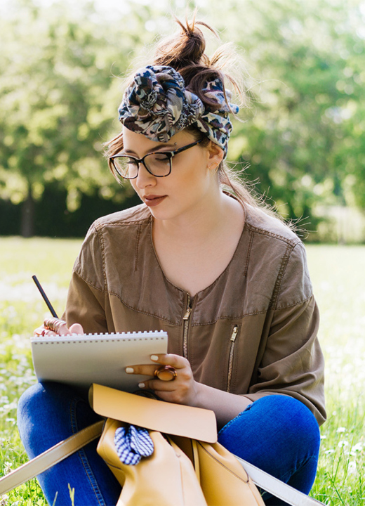 A young woman sitting in the grass, writing in a notebook surrounded by small white wildflowers and green trees.