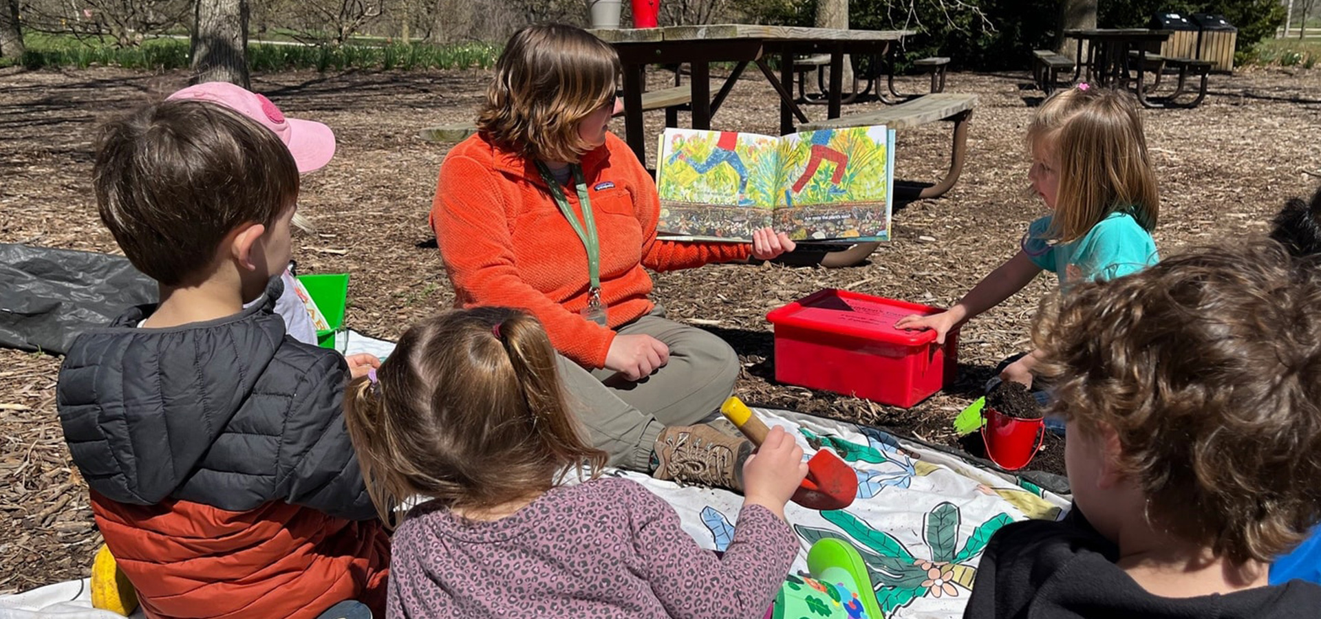 A photo of an Arboretum educator reading a book to a children's group outdoors at a picnic area at The Morton Arboretum