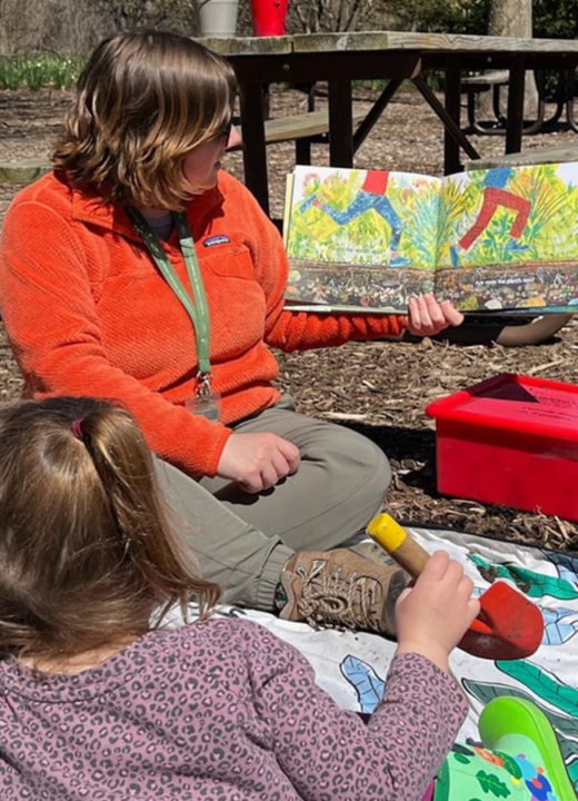 A photo of an Arboretum educator reading a book to a children's group outdoors at a picnic area at The Morton Arboretum