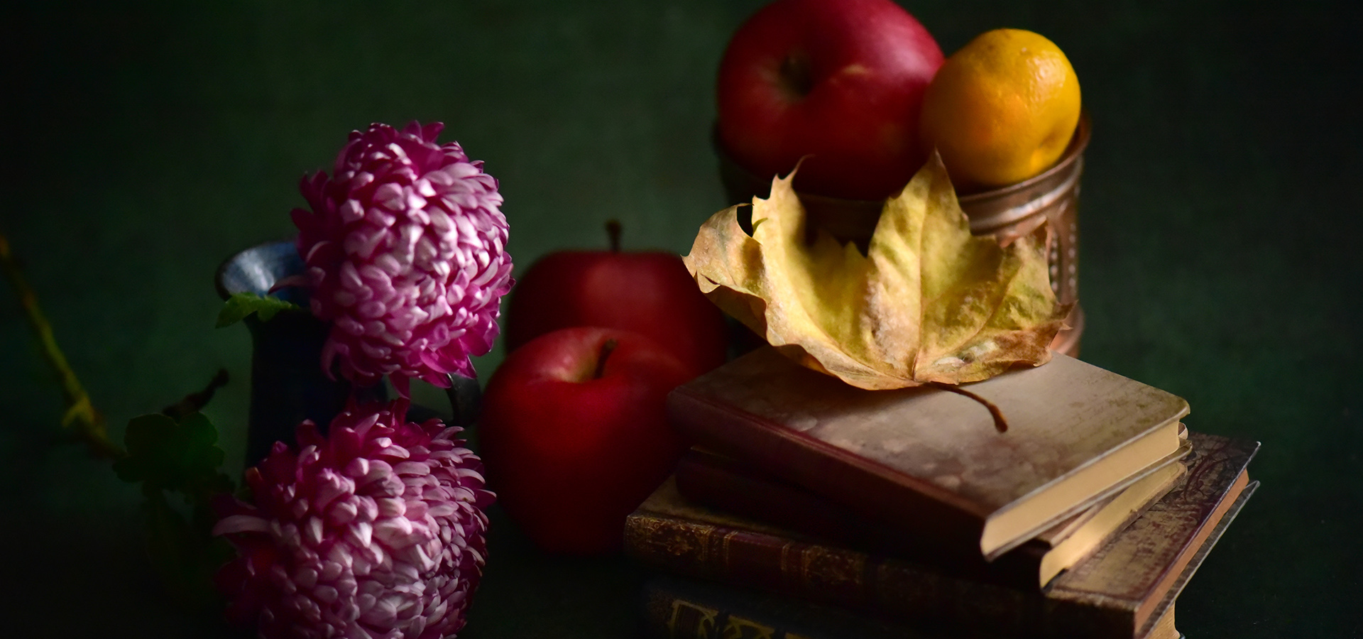 A still life photograph of books, a dried leaf, fruit, and flowers