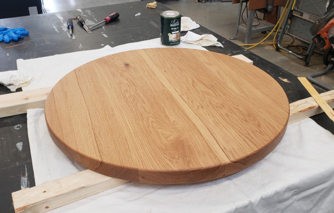 Wood stain is applied to round tabletop in woodworking shop