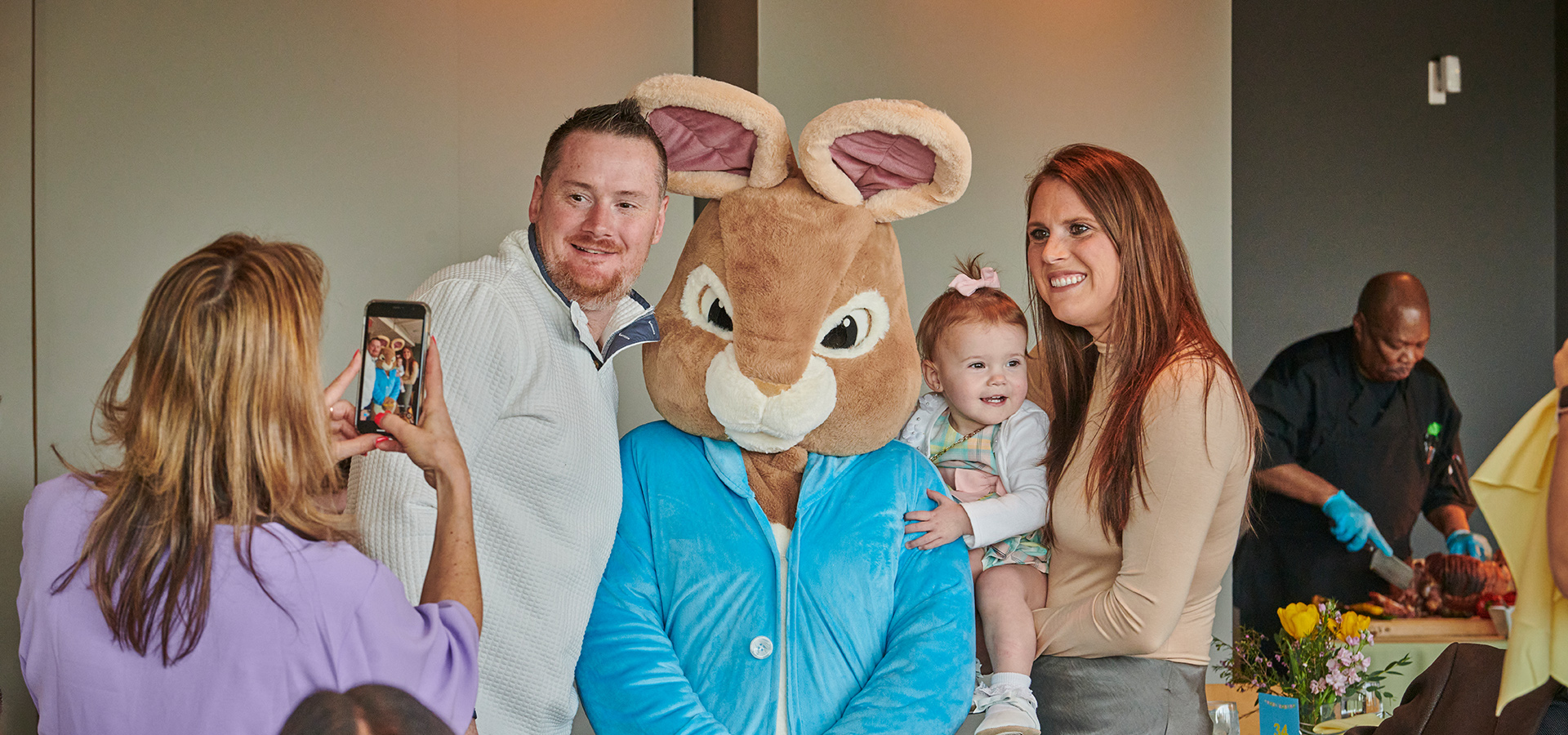 A family posing with the Easter Bunny while someone takes a photo with their phone in the Ginkgo restaurant during the Breakfast with the Bunny event.