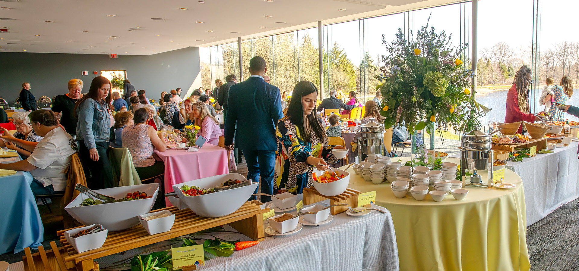 Visitors in a buffet line and sitting at tables in the Ginkgo restaurant. The restaurant is decorated with flowers and pastel colors for Easter.