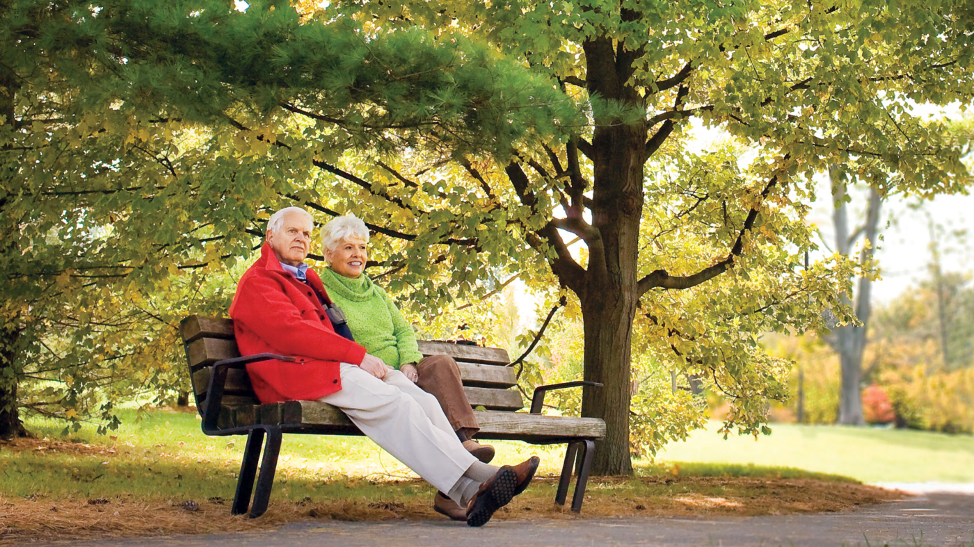 Photograph of grandparents sitting on a bench along a path on a sunny fall day