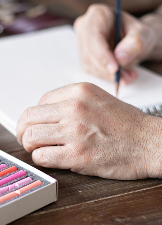 A person drawing in a sketchbook at a table full of colorful art supplies.