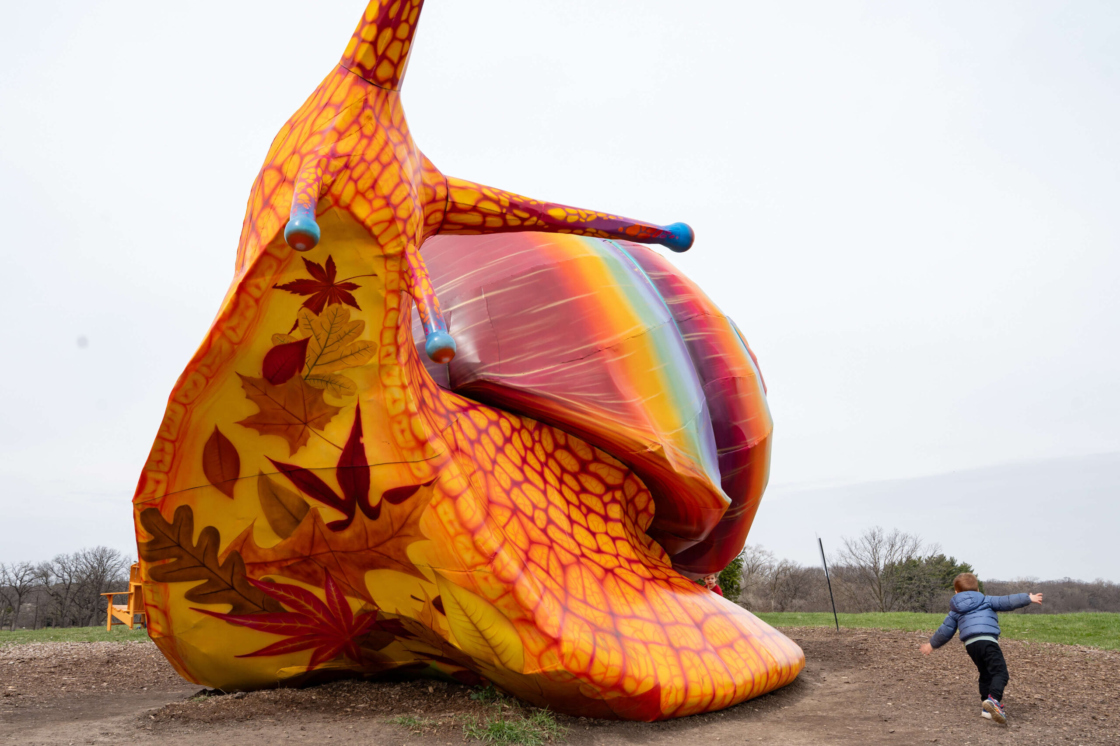 Small child races in a circle around larger-than-life snail sculpture; the angle reveals the snail's underside, which is painted with leaves in autumn colors