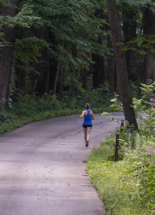 Runner on a paved road surrounded by tall green trees in summer