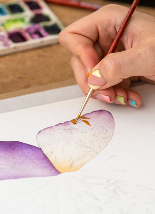 Artist uses a fine brush to add decorative detail to a painting of a butterfly using watercolors.