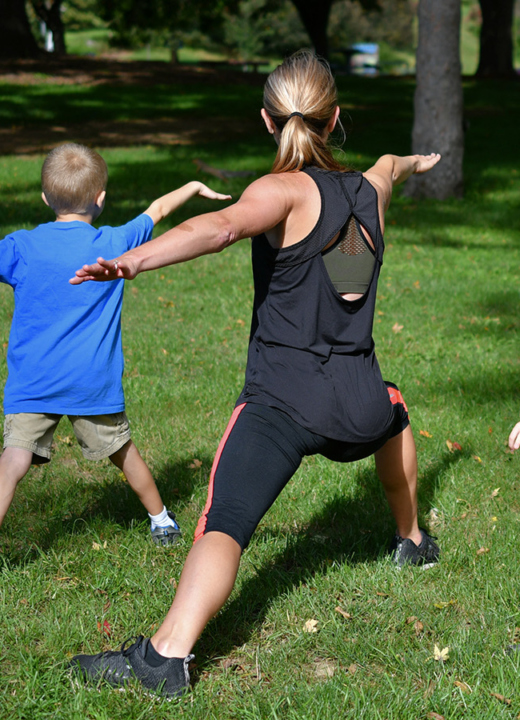 Yoga instructor demonstrates a pose for two young boys in a grassy area with trees in the background.