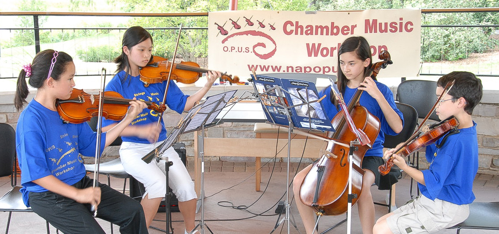 Four young musicians from OPUS Chamber Music Camp seated in front of music stands, performing outdoors as a string quartet.
