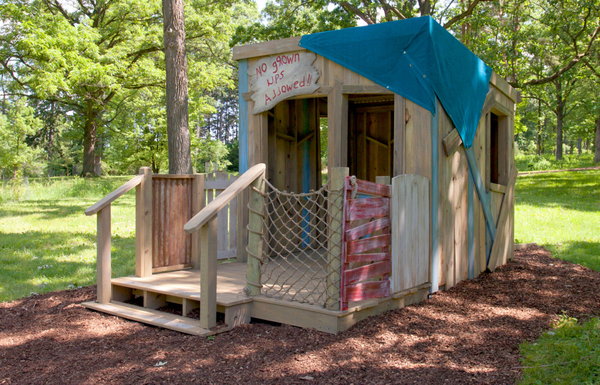 Makeshift shack built on grass in a wooded area, with sign in a child's handwriting that reads,