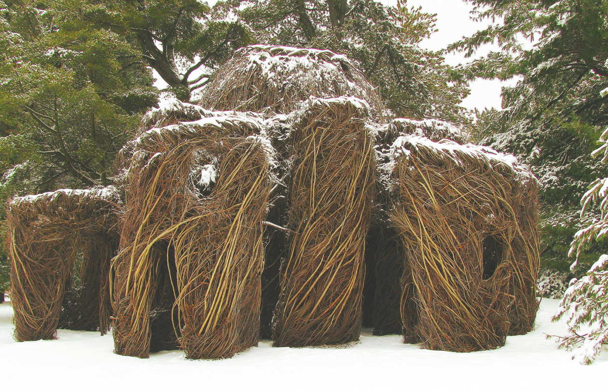 Xanadu, sculpture by American environmental artist Patrick Dougherty, a cluster of small shelters with windows and doorways, built from sticks and saplings, in a winter wooded landscape, frosted with snow.