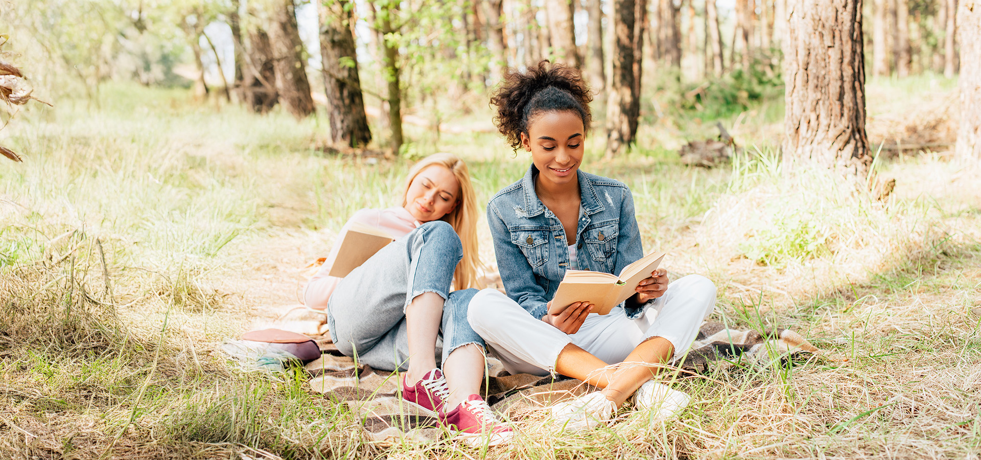 Two multiethnic women sitting on plaid blanket under trees reading books together