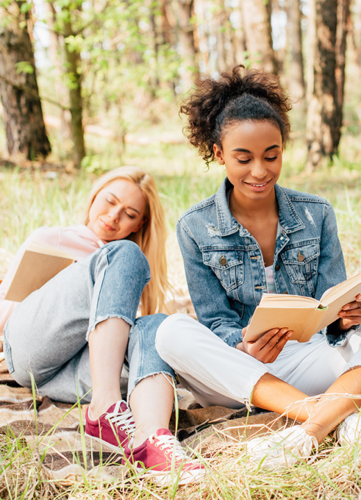 Two multiethnic women sitting on plaid blanket under trees reading books together