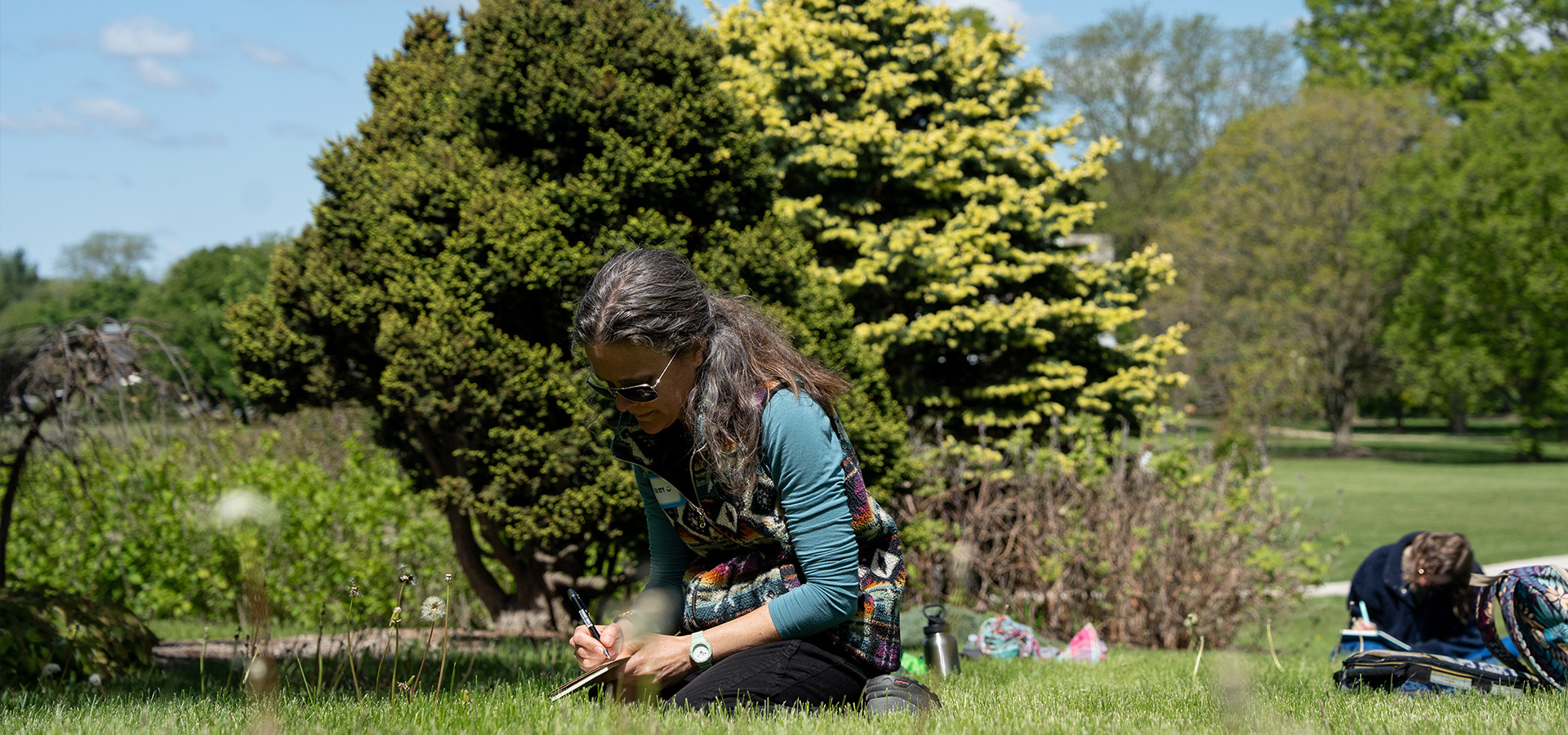 Photo of a woman kneeling on the grass at The Morton Arboretum writing in a journal