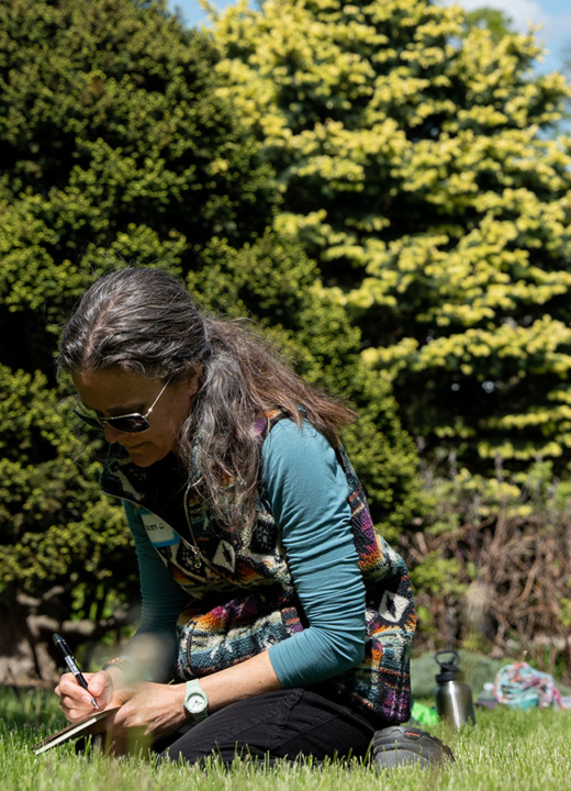 Photo of a woman kneeling on the grass at The Morton Arboretum writing in a journal