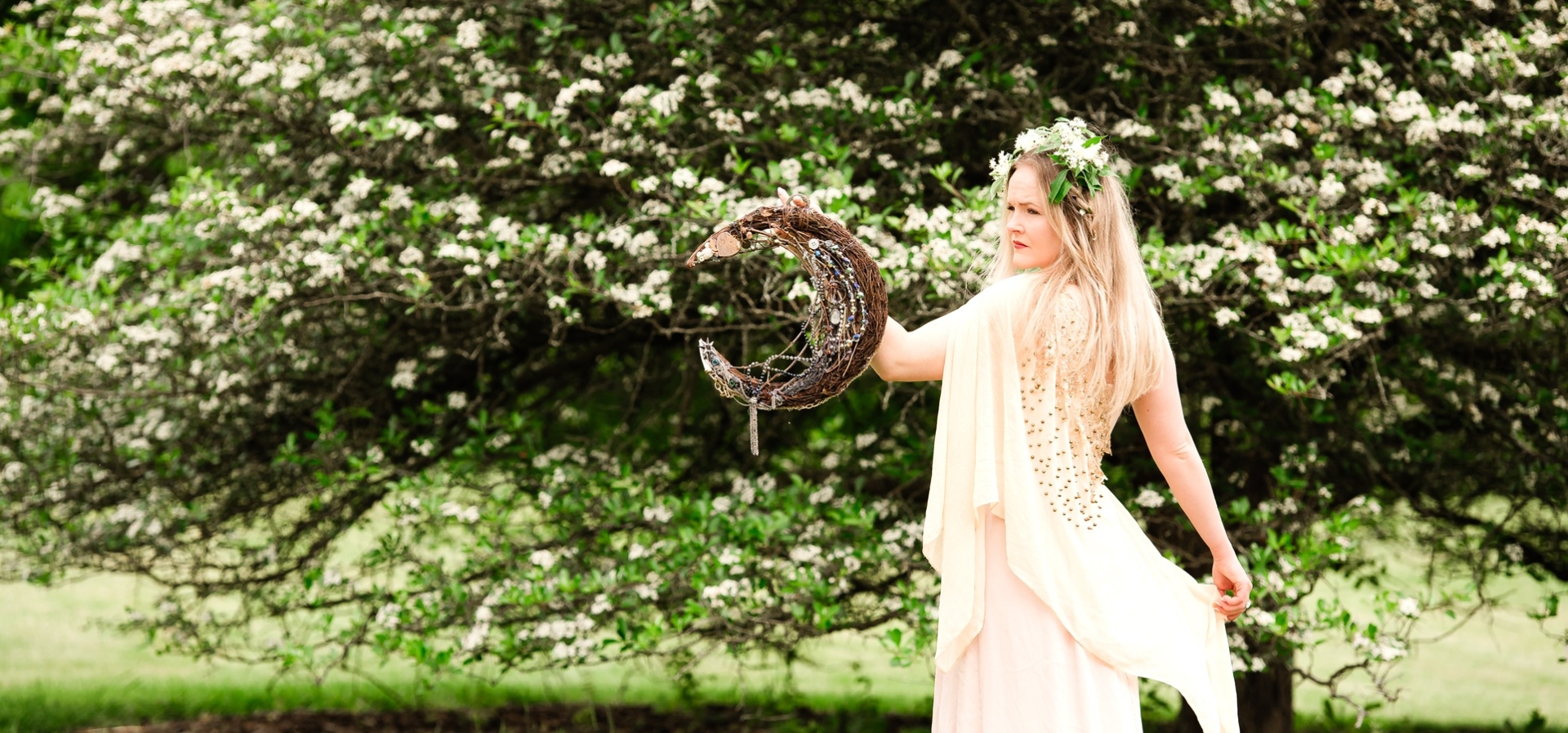Actor standing next to a flowering tree, wearing crown of branches and a flowing white dress, posing with a crescent moon prop made from sticks and beads