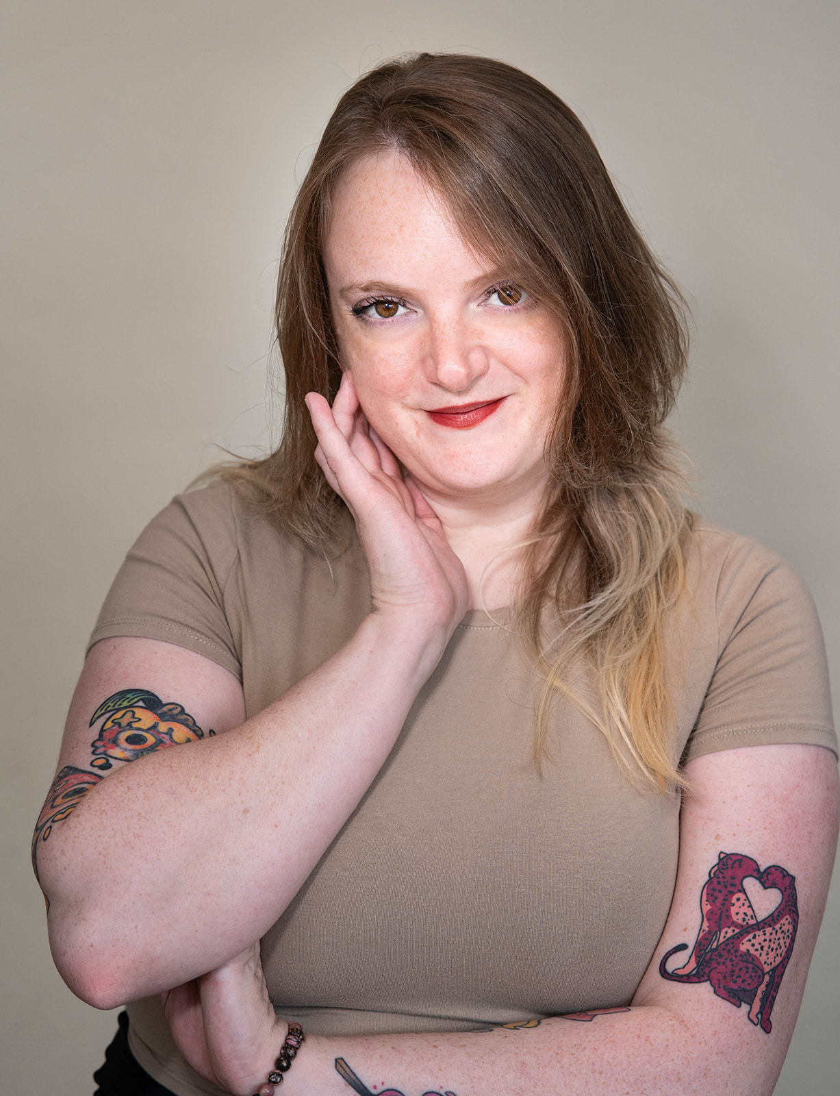 Headshot of woman with long brown hair wearing a tan T-shirt, with tattoos on both arms, including one of two red-and-orange cheetahs touching noses to form the shape of a heart between them