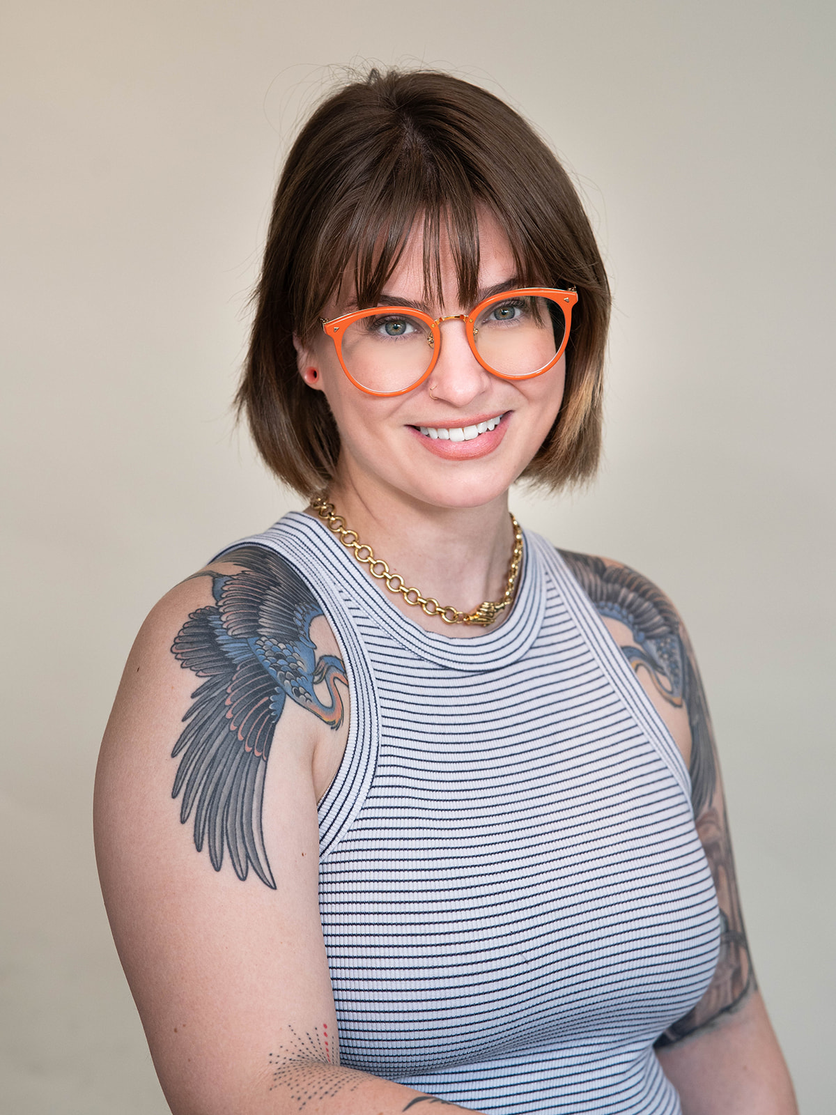 Headshot of a woman with chin-length brown hair, wearing oversized glasses with orange frames and striped tank top, and tattoos of outstretched bird wings on each shoulder