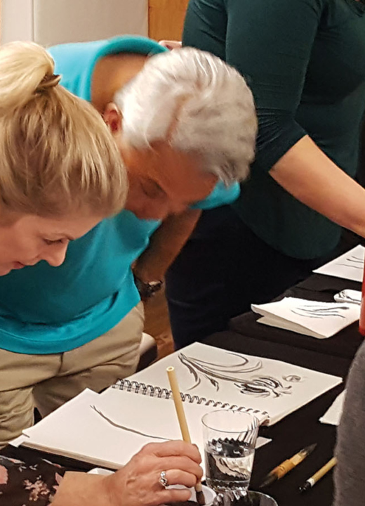 Artists stand at a table and use brushes to paint flowers in Sumi-e ink style