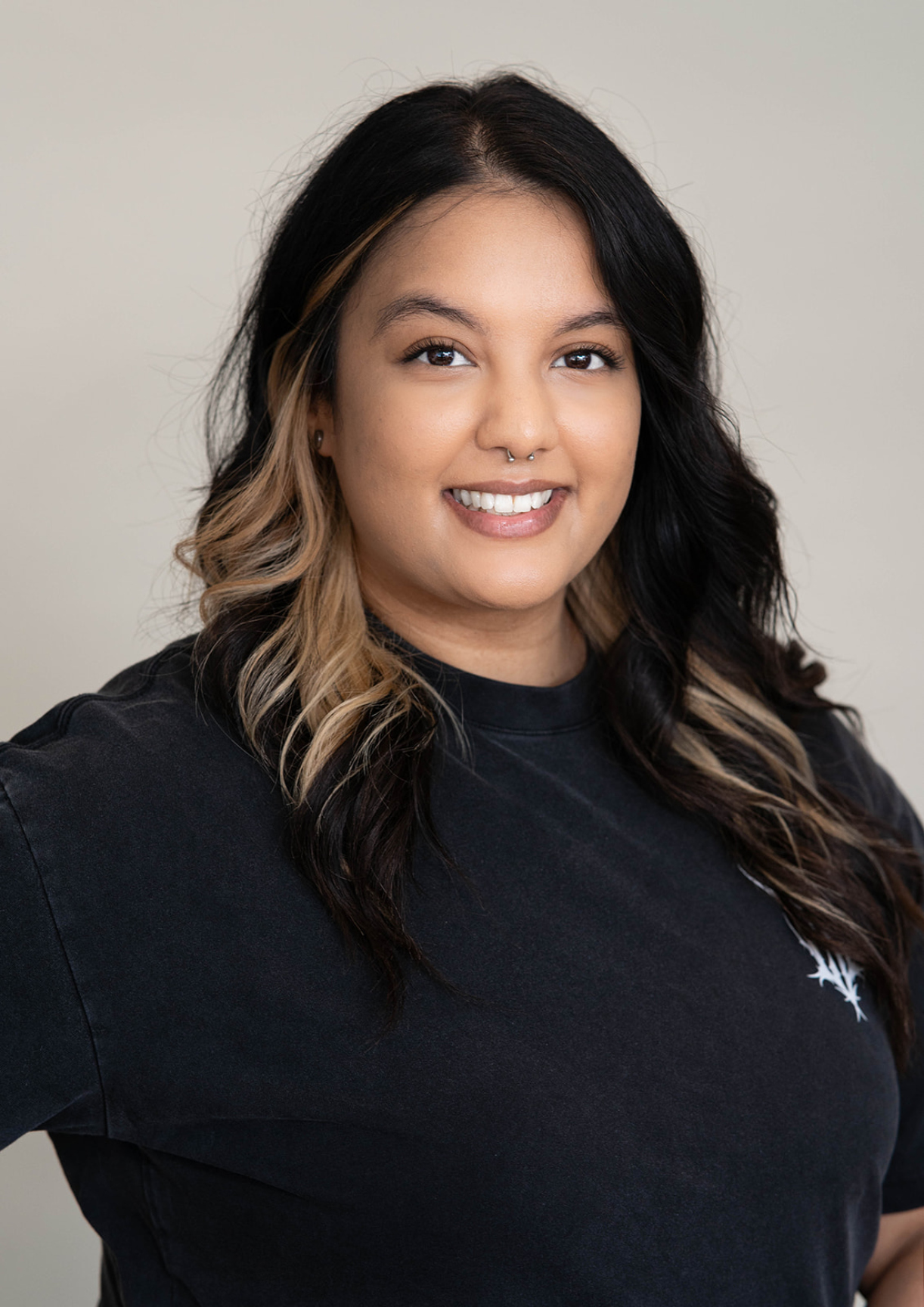 Headshot of woman with nose piercing and long hair, wearing a simple black T-shirt with small white design in the upper left corner