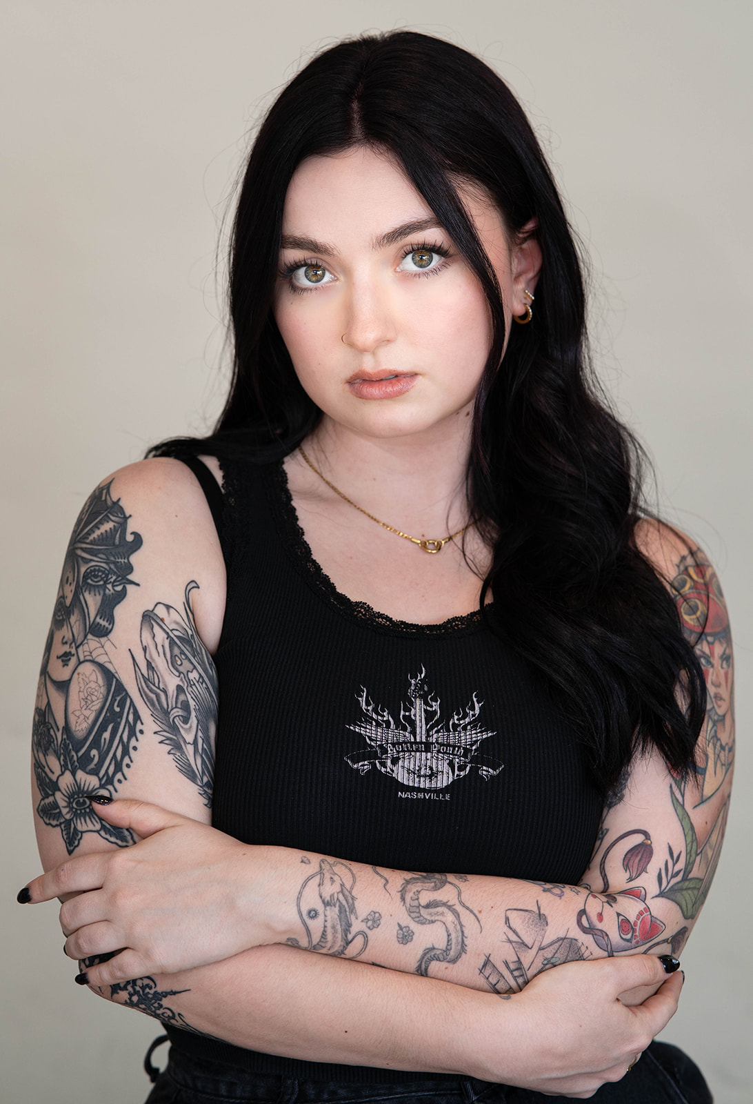 Headshot of woman with long black hair and hazel eyes, wearing a black tank top with small white design on front, with numerous black-and-white tattoos on her right arm and color tattoos on her left