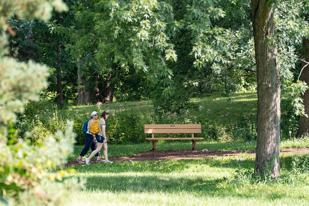 Two people hike past a tribute bench installed along a trail.